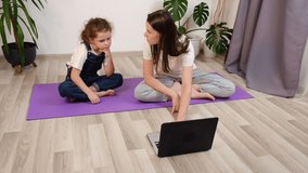 Young mom with little daughter sits on mat in living room in front of laptop watch online training class repeat exercises. Family choose what yoga exercise to do, look at laptop online class at home