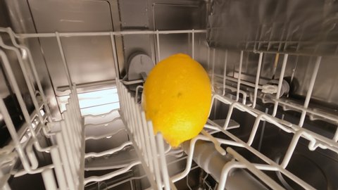 A stream of clean water washes the yellow lemon in the basket. Lemon in the white basket of the dishwasher. An idea for facilitating household management. Dishwasher, kitchen. High quality.