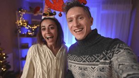 POV first person view happy cheerful excited couple, woman in festive Elf Cap and man in red reindeer antlers headband taking selfie on background of decorated house with christmas tree