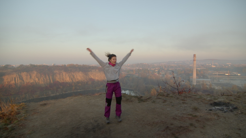 Young woman with curly hair dressed in sport clothes exercising on the top of the hill. City and mountain view on the background.