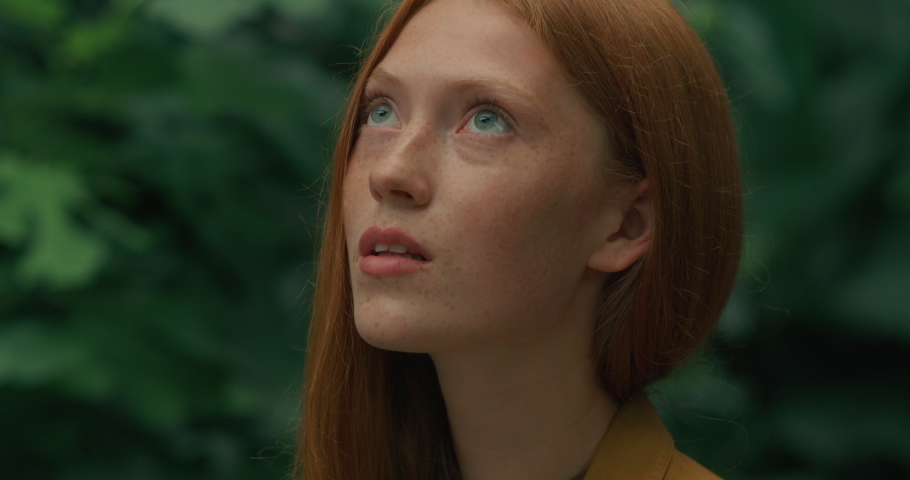 Portrait of Red-Haired Girl with Blue Eyes  on background of green plants. Ginger girl with freckles, looking up standing outside