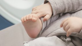 Baby boy hand holding his little bare foot, baby's leg in the hand, 8 month old caucasian kid in a rocking chair for babies close up. High quality 4k footage