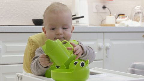 Cute kid in green bib holding baby drinker, 8 month old caucasian baby boy on feeding high chair, teaching infant to self feed. High quality 4k footage