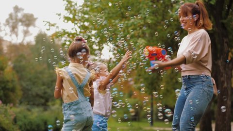 Happy mother blowing soap bubbles with toy gun, little daughters trying to catch bubbles flying in air. Family playing together, have fun in park. Mother's Day Joy, Mom and Baby