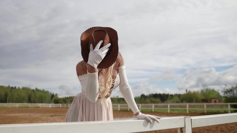 a blonde with long hair and in a revealing dress leaned hand on the fence and takes off cowboy hat, then puts it back on
