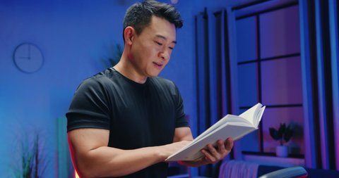 Close up of likable positive athletic 30-aged asian man in sportswear which reading interesting book during exercising on running track at home in the evening