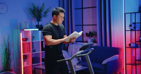 Attractive confident sporty modern asian guy in sportswear walking on treadmill and reading interesting book during evening home workout,slow motion