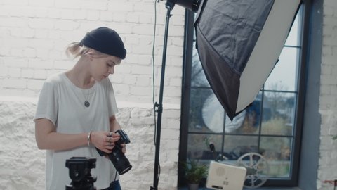 Isolated Caucasian Photographer Standing in Her Modern Photo Studio Holding a Camera and Setting Up Ready For the Photoshoot With a Beautiful  Caucasian Model. Wearing a Black Beanie and a White Shirt