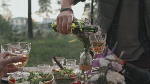 Close up mid-section of unrecognizable man pouring wine into glasses held by his friends sitting around table outdoors on summer day. People having picnic outside