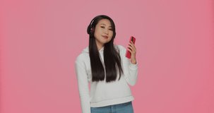 Asian girl listening music on phone in headphones having fun dancing using mobile musical apps on pink background