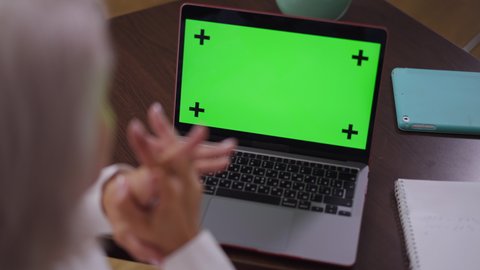 Close-up chromakey laptop on table in office with blurred mature businesswoman sitting indoors. Shooting over shoulder of middle aged Caucasian businesswoman thinking analyzing e-market. Mockup