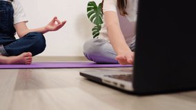 Home sports concept. Selective focus of young healthy mother and little child daughter doing yoga or meditation while watching online tutorial on laptop sitting together on purple mat in living room