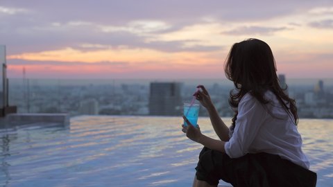Asian woman resting after work sitting on the ledge of the pool and enjoying refreshing cocktail in a glass with ice and a slice of lime on Blurred background city and Beautiful sunset sky. 