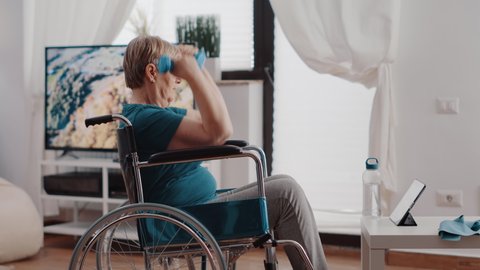 Aged person sitting in wheelchair using dumbbells to exercise while following online training lesson on tablet. Old woman with disability lifting weights and watching workout video.