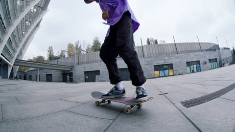 Bottom view of skater guy riding skateboard on urban background. Focused hipster making kickflip with skate outdoor in slow motion. Sporty skateboarder practicing in extreme sports outside in summer.