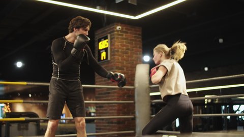 Closeup joyful opponents boxing at gym. Smiling male and female boxers practicing techniques in sport club. Friendly trainer teaching fit girl on boxing ring.