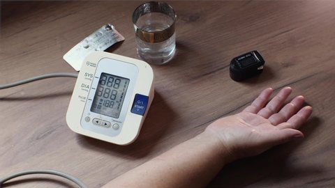 An elderly woman measures her blood pressure on an automatic blood pressure monitor. On a wooden table there is a hand, the device, a glass of water, and pills. Krasnodar, Russia September 12, 2021