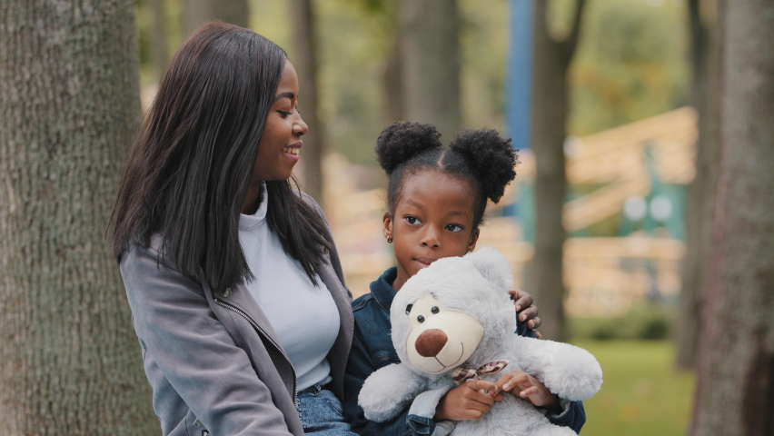 Joyful mom and daughter sitting on bench in park young mother hugs child shy little girl holding teddy bear african american family fun chatting outdoors mommy happily smiling kid spend time with mum Royalty-Free Stock Footage #1083280078