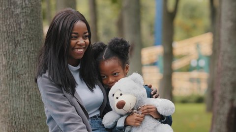 Joyful mom and daughter sitting on bench in park young mother hugs child shy little girl holding teddy bear african american family fun chatting outdoors mommy happily smiling kid spend time with mum