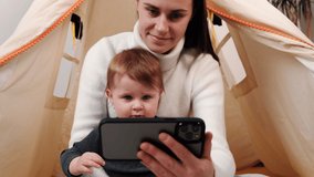 Addicted to modern technology concept. Close up happy mum and cute baby daughter having fun using cellphone. Young nanny playing mobile games with adorable small toddler girl sits at cozy tent at home