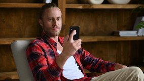 Online video communication. Positive friendly American young adult man, sitting on armchair at home, uses a cellphone, talks on a video call with friend or family, chatting on social networks, smiles