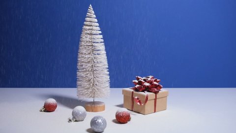Beautiful Christmas or New Year video banner with copy space. Toy Christmas tree, gift box with red bow and Christmas baubles on blue background with snow falling on the background.