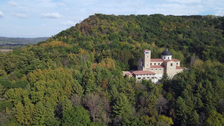 Aerial view of mountains in Wisconsin showing shrine peaking through the trees. Foliage on mountain changing to brilliant fall colors. Peaceful church in the woods on a hill with blue sky. 