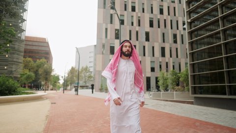 Successful arab man sheikh in traditional clothes looking at skyscrapers while walking city street