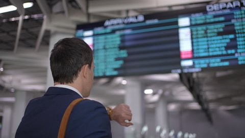Young man traveller in airport near flight timetable. Businessman in jacket with luggage at airport terminal arrivals table. Waiting for flight delay plane In terminal, travel business concept