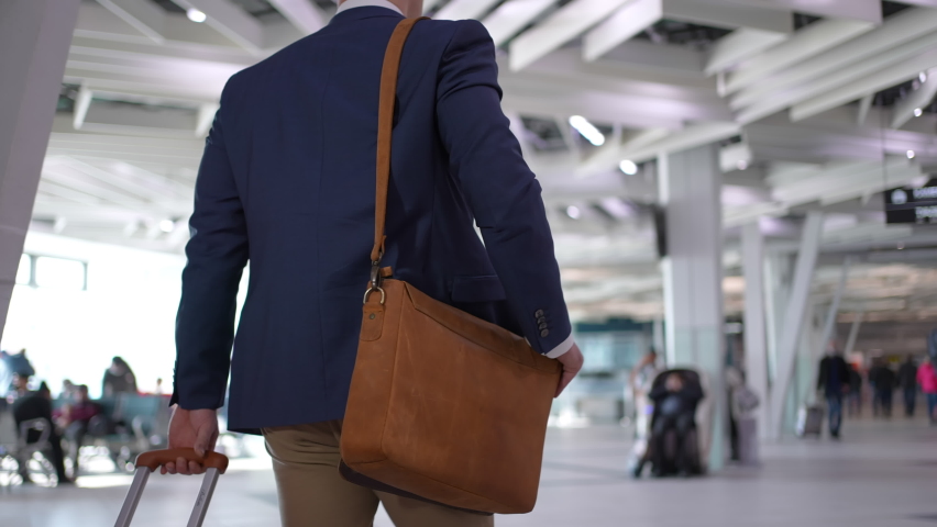 Businessman passenger pulling suitcase, handbag luggage in modern airport terminal. Young traveler man carries shoulder bag walking in airport, close-up rear view, Travel business concept Royalty-Free Stock Footage #1083290497