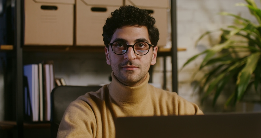 Young curly haired brunette man with stubble on his face takes off his glasses, and smiling while looks at the camera while sitting at the table in front of an open laptop in the office