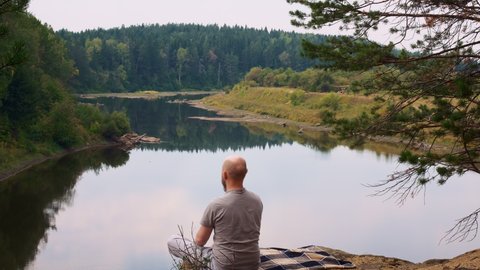 A bald man with a beard sits on the edge of a river bank cliff and meditates in a calm environment in nature in the forest and is filled with the energy of the earth. Yoga in nature and relocation.