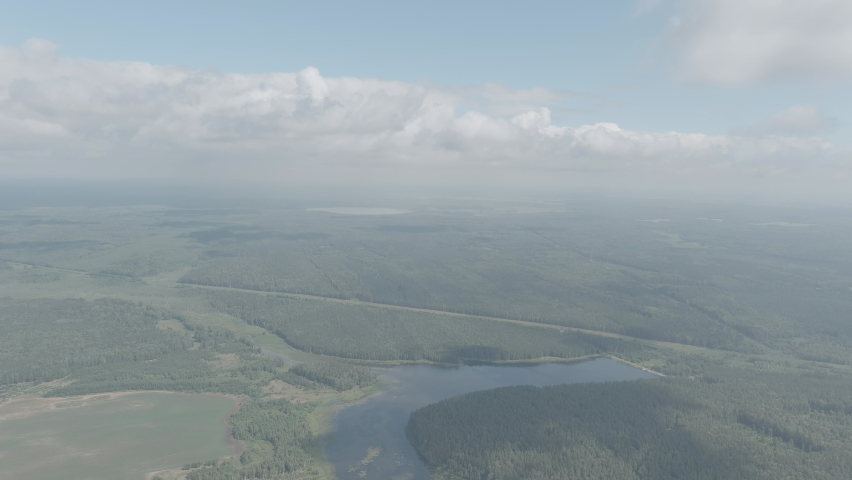 Russia, Ural. Aerial footage captures a sweeping high-altitude view over expansive boreal forest, patchwork agricultural fields, and a winding river that cuts through the wooded landscape. A straight road runs parallel to the tree line, dividing cultivated land from dense woodland. Cloud shadows drift across the terrain, creating dynamic patterns of light and shade on the green canopy below. The horizon stretches into a soft atmospheric haze under a partly cloudy summer sky. The camera maintains a steady forward glide, offering a broad panoramic perspective of this remote rural region. The scene conveys the vast scale and natural tranquility of the Ural countryside, with no visible human activity beyond the road and scattered clearings. This footage is suited for travel, nature documentary, and environmental stock use. A-Log.