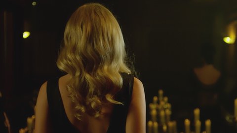 Back view of beautiful blond woman in black elegant dress  walking slowly in dark room with lighted candles . Model walking in luxury fashion show . Expensive home interior . Slow motion .