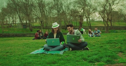 Beautiful young girl sitting on grass or lawn in the park and working on laptop . Boy coming asking to sit . Male and female students, friends talking , studying and discussing together . Slow motion
