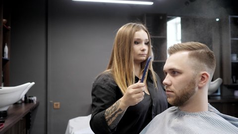 Young woman hairdresser with long blond hair and tattoo on her arm combing hair to client brutal man with beard: Gomel, Belarus - July 2020