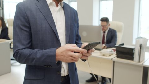 Tracking mid-section close up of unrecognizable businessman in suit walking through office and typing on mobile phone