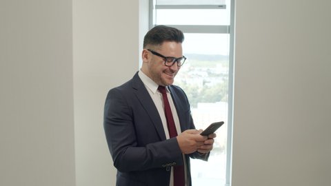 Tracking shot of successful male CEO in suit and glasses standing by window in office and laughing while typing on mobile phone