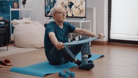 Retired adult pulling resistance band and sitting on yoga mat at home. Mature person doing stretching exercise with flexible elastic belt to train arms muscles. Pensioner training