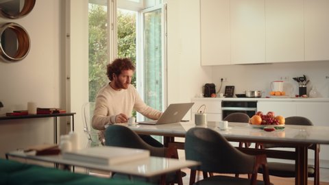 Handsome Adult Man with Ginger Curly Hair Using Laptop Computer, Sitting in Living Room in Apartment. Male Drinks Espresso, Working from Home, Online Shopping, Watching Videos or Writing Emails.