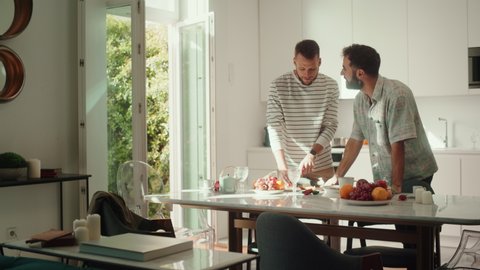 Stylish Young Adult Gay Couple at Home in Casual Clothes in Kitchen Area. Handsome Male Cutting Apples and Preparing Lunch. Partner Comes Over and Gives a Kiss. Cute LGBTQ Relationship Content.