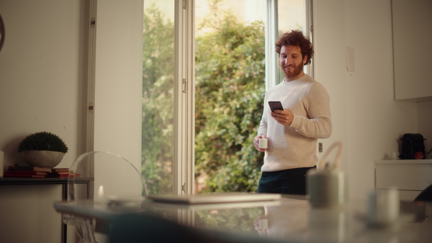 Handsome Adult Man with Ginger Curly Hair Using Smartphone, Standing in Living Room at Home. Male Enjoys a Cup of Espresso, Checking Social Media, Online Shopping, Watching Videos or Writing Messages. | Shutterstock HD Video #1083304450