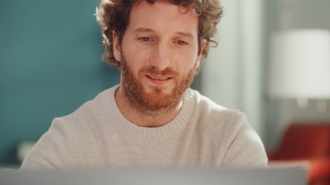 Close Up Portrait of a Handsome Adult Man with Ginger Curly Hair Using Laptop Computer, Sitting in Living Room. Smart Man is Working from Home, Online Shopping, Watching Videos or Writing Emails.