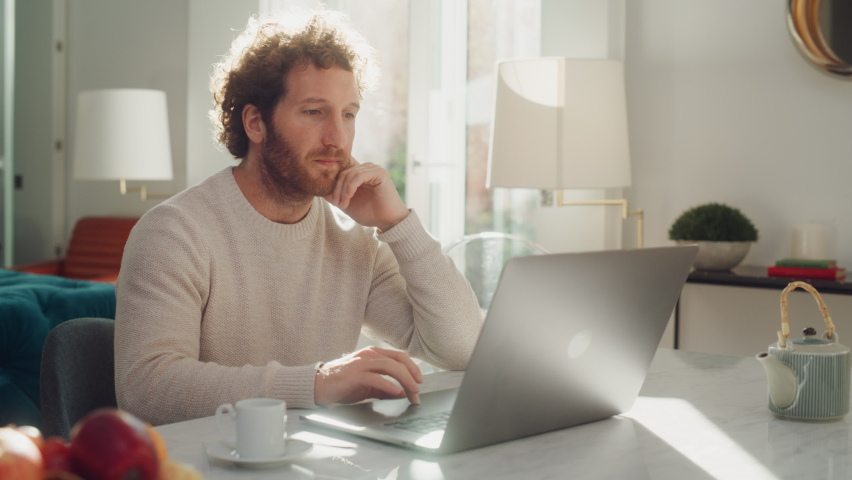 Handsome Adult Man with Ginger Curly Hair Using Laptop Computer, Sitting in Living Room in Apartment. Attractive Man is Working from Home, Online Shopping, Watching Videos or Writing Emails. Royalty-Free Stock Footage #1083304480