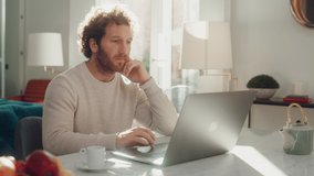Handsome Adult Man with Ginger Curly Hair Using Laptop Computer, Sitting in Living Room in Apartment. Attractive Man is Working from Home, Online Shopping, Watching Videos or Writing Emails.