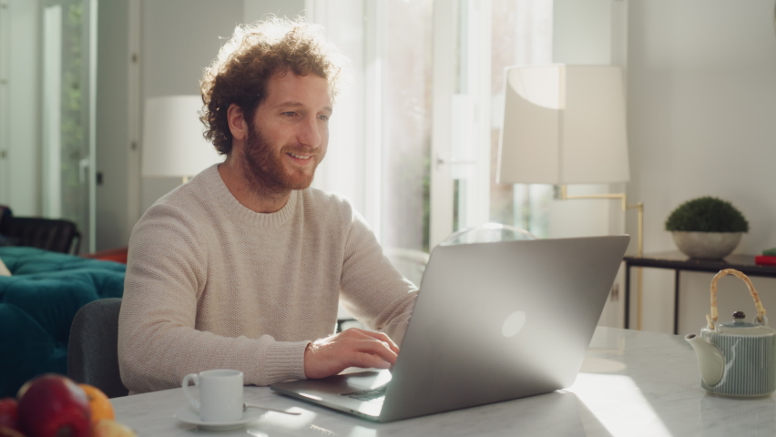 Handsome Adult Man with Ginger Curly Hair Using Laptop Computer, Sitting in Living Room in Apartment. Attractive Man is Working from Home, Online Shopping, Watching Videos or Writing Emails.