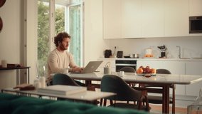 Handsome Adult Man with Ginger Curly Hair Using Laptop Computer, Sitting in Living Room in Apartment. Male Drinks Espresso, Working from Home, Online Shopping, Watching Videos or Writing Emails.