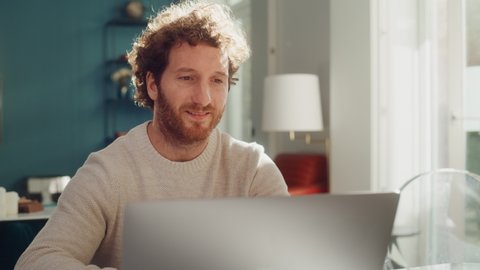 Handsome Adult Man with Ginger Curly Hair Using Laptop Computer, Sitting in Living Room in Apartment. Dedicated Man is Working from Home, Online Shopping, Watching Videos or Writing Emails.