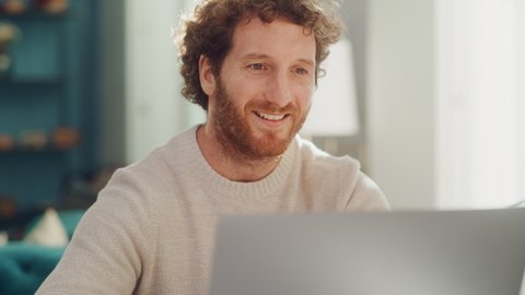 Handsome Adult Man Portrait with Ginger Curly Hair Using Laptop Computer, Sitting in Living Room in Apartment. Joyful Man is Working from Home, Online Shopping, Watching Videos or Writing Emails.