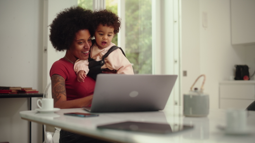 Modern Young Mom Holding Her Adorable Baby Son, Using Laptop Computer in Living Room in Apartment. Mother is Working from Home, Online Shopping, Watching Social Media or Writing Emails. | Shutterstock HD Video #1083306841